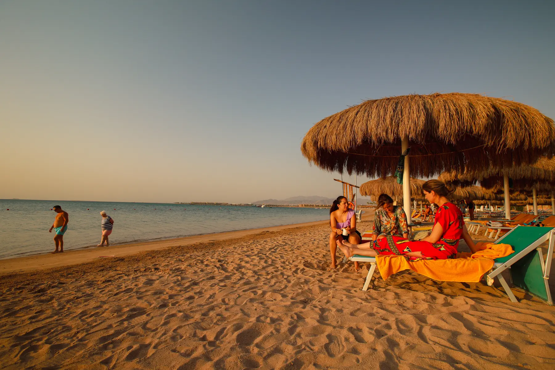 Guests relaxing with coffee in the beach lounge area while others walk along the shoreline at Caribbean World Resorts Hurghada